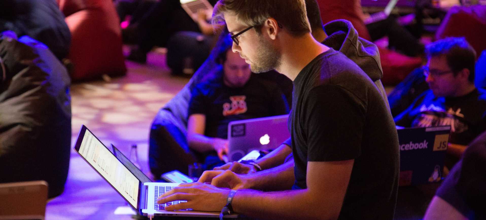 A man with glasses in a group of people on a laptop in a colorfully lit general session